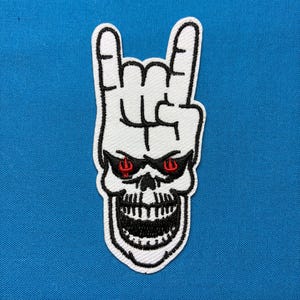 Könnte beinhalten: Ein gestickter Aufnäher mit einer weißen Hand, die ein Rock-and-Roll-Zeichen formt, über einem weißen Totenkopf mit schwarzen Umrissen und roten Augen. Der Totenkopf hat einen weit geöffneten Mund mit sichtbaren Zähnen und ist weiß umrandet.