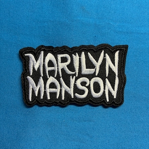 May include: A rectangular embroidered patch with a black background and white stitched text that reads MARILYN MANSON. The lettering features a jagged, stylized font with a thick black border and a textured surface finish throughout the design.