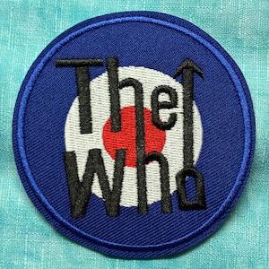 Puede incluir: Un parche bordado azul circular con el texto "The Who" en negro, con un diseño de diana blanco y rojo en el centro. El parche tiene un borde azul y está sobre un fondo azul claro texturizado.