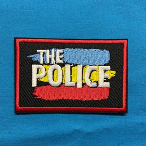 May include: A rectangular embroidered patch with a black background and a red border. The text THE POLICE is stitched in white, layered over three horizontal brushstroke-style stripes in light blue, yellow, and red, creating a bold graphic design.