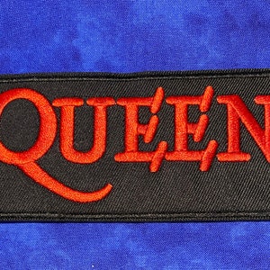 May include: Embroidered patch with the word "QUEEN" in red thread on a black rectangular background. The patch is set against a blue fabric backdrop.