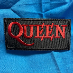 Peut inclure: Un écusson rectangulaire brodé présentant un fond noir avec le mot QUEEN cousu en fil rouge vif. Le lettrage est stylisé avec une queue proéminente et incurvée sur la lettre Q et des barres horizontales traversant les lettres U, E, E et N.