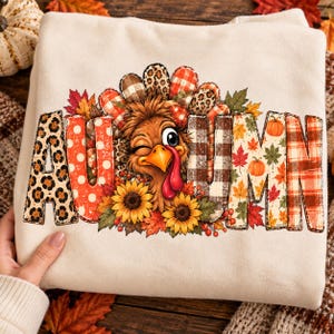 Könnte beinhalten: Ein cremefarbenes Sweatshirt mit einer Grafik eines zwinkernden Truthahns, umgeben von Sonnenblumen und Herbstblättern. Das Wort AUTUMN ist in großen Buchstaben gedruckt, die jeweils mit verschiedenen Mustern wie Leopardenprint, Punkten und Karos gefüllt sind.