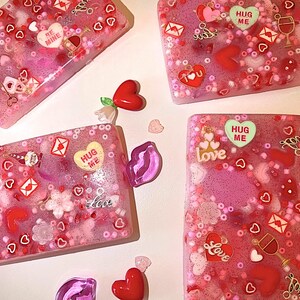 May include: Four rectangular, pink resin art pieces with embedded hearts, glitter, and decorative elements. Some pieces feature the words "Love", "Hug Me", and "Be Mine". Red heart-shaped embellishments and small heart confetti are scattered around the pieces.