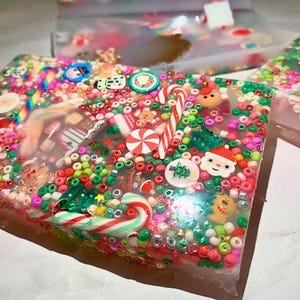 Könnte beinhalten: Rechteckiges Kunstwerk aus Harz mit weihnachtlichen Verzierungen. Das klare Harz umschließt bunte Perlen, Zuckerstangen, Weihnachtsmann-Gesichter und Weihnachtsbaum-Designs. Das Stück ist mit roten, grünen, rosa und weißen Elementen gefüllt.