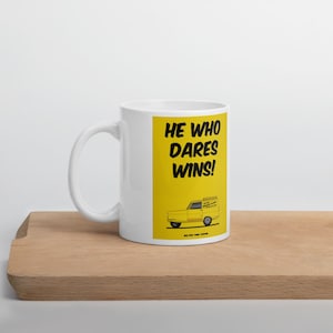 May include: White ceramic mug with a yellow panel featuring the text "HE WHO DARES WINS!" and a yellow van illustration. The mug is on a wooden surface.