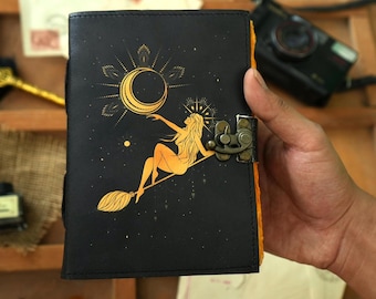 Puede incluir: Un diario de cuero negro con un diseño dorado de una mujer volando en una escoba con una luna creciente y estrellas. El diario tiene un cierre de latón.