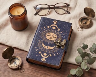 Puede incluir: Un diario azul oscuro con una ilustración dorada de sol y luna en la cubierta, asegurado con un cierre de metal. El diario está rodeado por una vela en vaso marrón, unas gafas de montura oscura, dos brújulas de latón y una rama de hojas.