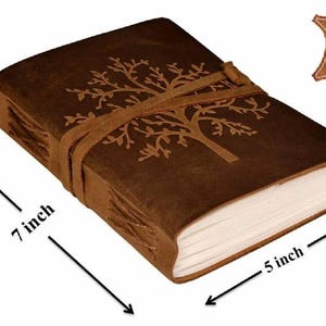 May include: A brown leather journal featuring an embossed tree design on the cover, secured with a leather wrap tie. The journal measures 7 inches by 5 inches and includes a 100% Genuine Leather label in the corner, with visible side stitching.
