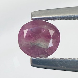May include: A faceted, oval-shaped ruby gemstone held by a pair of silver tweezers. The ruby displays a deep pink color with lighter pink and white inclusions. The gem is cut with multiple facets to reflect light.