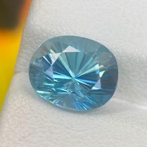 May include: A faceted, oval-shaped gemstone with a light blue hue. The gem's surface is intricately cut, creating a starburst pattern emanating from the center. The gem is set against a white background.