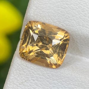 May include: A faceted, cushion-cut gemstone with a warm, golden-yellow hue. The gem displays internal inclusions and a brilliant, reflective surface. The stone is set against a white background, with a blurred green and yellow backdrop.