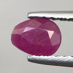 Puede incluir: Una piedra preciosa de rubí rosa con un corte facetado. El rubí se sostiene con unas pinzas.