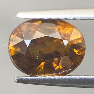 May include: A brown oval-shaped gemstone with a faceted cut. The gemstone has a golden hue with a slight green tint.