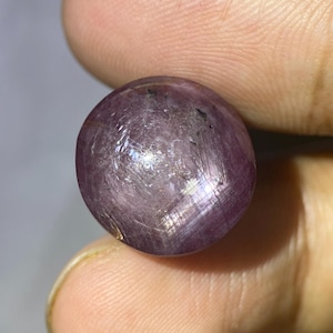 May include: A polished, round, purplish-red gemstone held between fingers. The gem has a smooth, reflective surface with visible internal striations and small dark inclusions. The overall appearance is a deep, rich color.