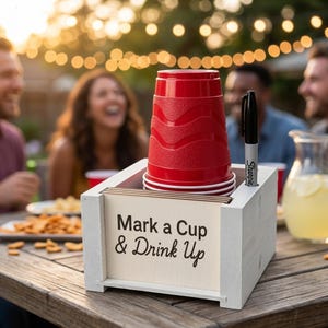 May include: A white wooden square holder containing a stack of red plastic cups and a black marker. The front panel features the black printed text "Mark a Cup & Drink Up". The holder is designed to store cups and a writing utensil for party use.