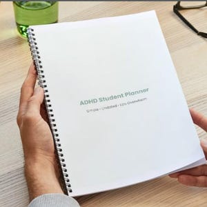 Puede incluir: Un planificador de estudiantes con TDAH encuadernado en espiral blanco, con el texto "Simple - Sin fecha - Menos agobio". El planificador está abierto, revelando páginas en blanco. Un vaso de líquido verde y gafas son visibles en el fondo.