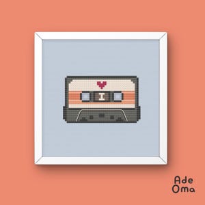 May include: A framed cross-stitch of a retro cassette tape design. The tape is in shades of gray, beige, and orange, with a red heart symbol. The artwork is set against a light blue background within a white frame.
