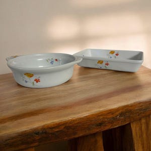 May include: Two white ceramic baking dishes with floral designs. The smaller dish is round with two handles and the larger dish is rectangular. Both dishes have a white background with small yellow, orange, and blue flowers.