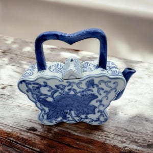 Puede incluir: Una tetera pequeña de porcelana azul y blanca con forma de mariposa. La tetera tiene un asa azul y una tapa con un pequeño pico. La tetera está decorada con un diseño floral.