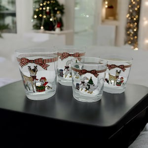 May include: A set of four clear glass tumblers with festive Christmas designs. Each glass has a red plaid bow detail and illustrations of deer, geese, and presents. The glasses are displayed on a dark surface, perfect for seasonal drinks.