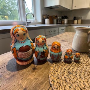 Könnte beinhalten: Ein Set aus fünf russischen Matrjoschka-Puppen in verschiedenen Größen. Die Puppen zeigen bemalte Gesichter und traditionelle Kleidung in Blau-, Orange- und Rottönen. Die größte Puppe trägt den Text "Russian National Orchestra".