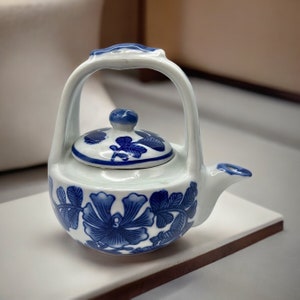 May include: A small, white porcelain teapot with a blue and white floral design. The teapot has a handle that forms a loop around the top of the teapot.
