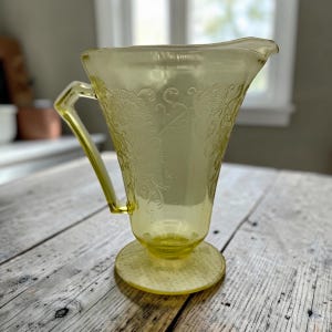 May include: A pale yellow translucent glass pitcher featuring an embossed floral pattern across its body. The vessel has a wide pouring spout, a sturdy angular handle, and a circular pedestal base, all crafted from a single piece of molded glass.