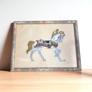 Può includere: Opera d'arte incorniciata raffigurante un cavallo bianco da giostra con criniera e coda dorate, adornato con una sella decorativa. Il cavallo è ambientato su uno sfondo beige, incorniciato in un bordo a motivi multicolori. Firmato in basso a destra.