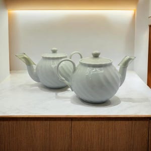 May include: Two white ceramic teapots with a swirl design. The teapots have a spout and a handle on each side.