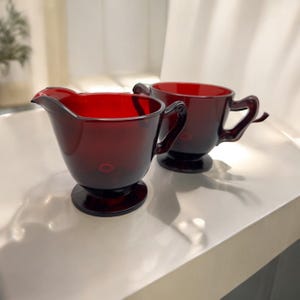 May include: A red glass creamer and sugar bowl set. The creamer has a rounded body and a handle. The sugar bowl has a rounded body and two handles.