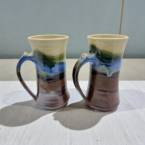 May include: Two handmade ceramic mugs with a unique, layered color design. The mugs feature a cream-colored rim, transitioning to green, blue, and brown bands. Each mug has a curved handle and a flared base, perfect for enjoying a warm beverage.