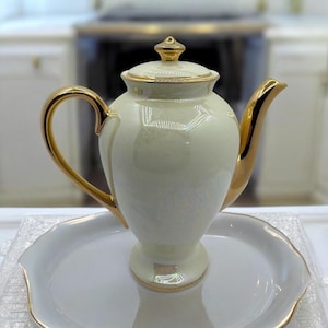 May include: Cream-colored porcelain teapot with gold accents on the handle, spout, and lid. The teapot sits on a matching white tray with a gold trim. The design is elegant and classic, suitable for serving tea.
