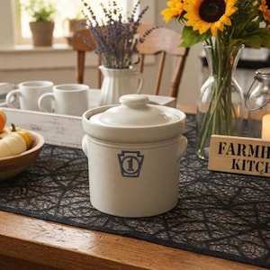 May include: A cream-colored ceramic crock with a lid, featuring a blue keystone-shaped logo with the number 1 in the center. The crock sits on a black patterned table runner next to a glass vase of sunflowers and white mugs.