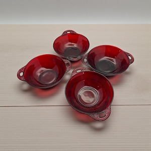 May include: Four ruby red glass bowls with handles. Each bowl has a round base and a flared rim with vertical ridges. The handles are ornate with a decorative design. The bowls are arranged on a light wood surface.
