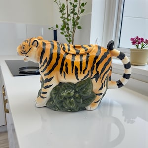 May include: A ceramic teapot shaped like a tiger, featuring orange and black stripes. The teapot has a green leaf base and a black and white striped handle. The lid is on top, and the teapot is on a white surface.