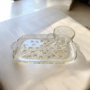 Può includere: Un set di tazza e piattino da tè in vetro trasparente. Il piattino rettangolare presenta un motivo decorativo a forma di goccia e manici su ciascuna estremità. La tazza da tè ha un manico e un motivo a rombi. Il set è su una superficie bianca.