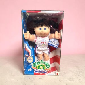 May include: A Cabbage Patch Kids Olympikids doll with dark hair and blue eyes, wearing a white and blue basketball uniform with USA and Olympic rings branding. The doll is inside a box featuring red, white, and blue star and stripe graphics.