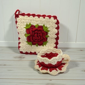 May include: A crocheted pot holder and teacup set. The pot holder is square, with a red rose in the center, surrounded by green leaves. The teacup and saucer are also crocheted, with a red and cream color scheme.