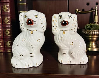 Puede incluir: Dos figuras de perro de cerámica blanca con detalles dorados. Los perros están sentados con las orejas levantadas, con narices negras y bocas rojas. Las figuras tienen un collar dorado y se exhiben en una estante marrón oscuro.