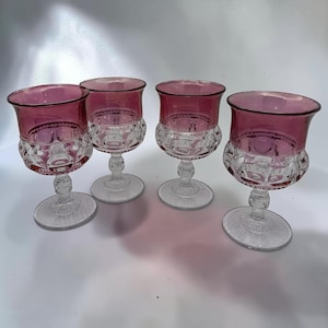 May include: A set of four stemmed goblets featuring a deep magenta-colored upper bowl and a clear glass base. The lower portion of each bowl displays a textured, circular pattern, while the stems and circular feet are made of clear glass.