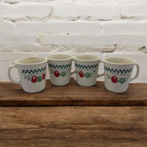 Può includere: Quattro tazze in ceramica bianca con bande a scacchi verdi e nere e un design di frutta verde e rossa. Le tazze recitano "Farm Fresh" e "Homemade".
