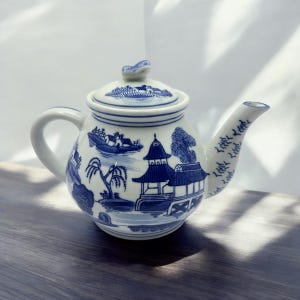 May include: A blue and white porcelain teapot with a traditional Chinese design. The teapot has a round body and a spout on the side. The design features a pagoda, trees, and water.