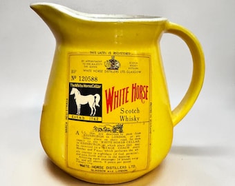 Op de afbeelding: Een vintage gele keramische kan met een handvat, met een "White Horse Scotch Whisky" label. Het label bevat tekst, een witte paardenillustratie en de tekst "Estab 1742".