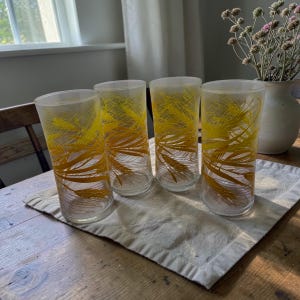Peut inclure: Quatre verres à boire transparents avec un motif de blé jaune. Les verres sont cylindriques et reposent sur une serviette en lin beige sur une table en bois. Un vase de fleurs séchées est à l'arrière-plan.