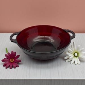 May include: A deep red glass bowl with two handles. The bowl has a ribbed texture and a dark rim. A dark red flower and a white flower with a yellow center are on the table.