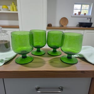 Op de afbeelding: Vier groene drinkglazen met een getextureerd oppervlak en een voet. De glazen staan op een houten aanrecht. De glazen hebben een afgeronde komvorm en een brede voet. De achtergrond toont een keuken met witte kasten en een raam.