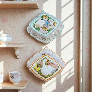 May include: Two decorative ceramic wall plaques. The top plaque features a lamb with a blue bow, surrounded by flowers and butterflies. The bottom plaque depicts a white goose with yellow butterflies and pink flowers. Both plaques have scalloped edges.