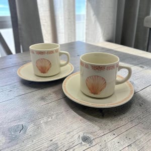 May include: Two off-white ceramic cups and matching saucers, each featuring a central illustration of a pinkish-orange scallop shell and a decorative border of small, repeating shell patterns in a soft coral hue around the rims.