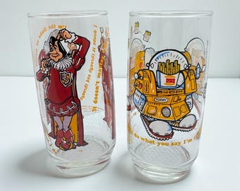 Puede incluir: Dos vasos transparentes con gráficos de dibujos animados. Un vaso presenta un personaje con un atuendo rojo y blanco con texto. El otro vaso tiene un personaje con forma de robot con papas fritas y el texto "YUMMY".